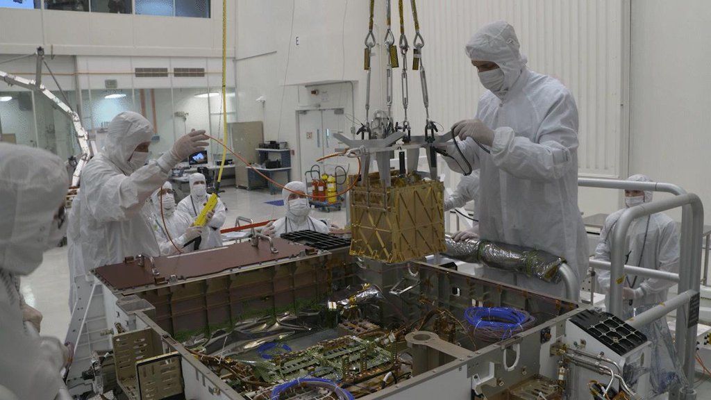 Engineers in cleanroom suits lower the gold MOXIE instrument into Perseverance’s chassis. NASA/JPL-Caltech, PIA24176.